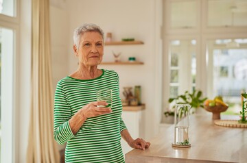 Ältere Frau in gestreiftem grünen Oberteil hält ein Glas Wasser in der Küche und blickt ruhig in die Kamera.